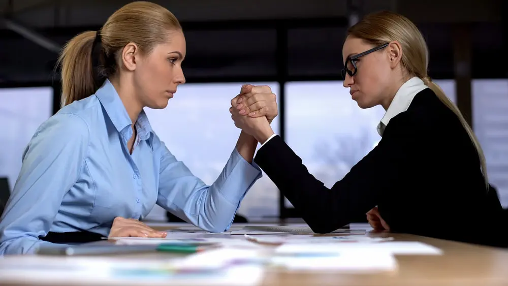 Two overly competitive woman coworkers arm wrestle at the office.