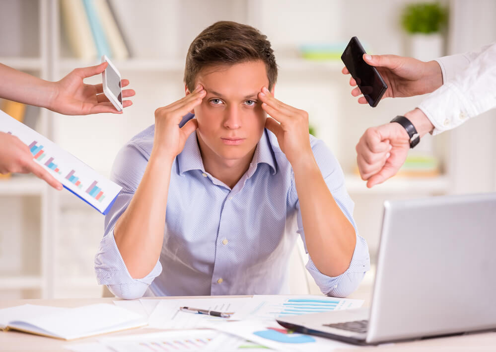 a frustrated worker rubs his temples while people thrust multitasking distractions at him