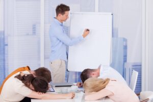 A meeting leader writes on a whiteboard and his team is asleep at the table.