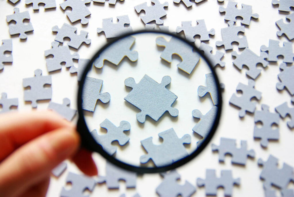 A man holds a magnifying glass on a tabletop full of puzzle pieces