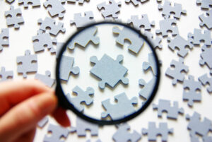 A man holds a magnifying glass on a tabletop full of puzzle pieces
